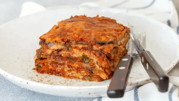 Porción de pastel de berenjenas, con capas de berenjena y salsa de tomate, servido en un plato blanco con cubiertos. ¡Un pastel de berenjenas facilisimo!