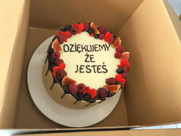 Okrągły tort z białym kremem, ozdobiony owocami i serduszkami. Krótki napis na tort urodzinowy: "DZIĘKUJEMY ŻE JESTEŚ".