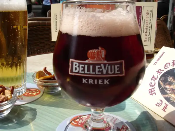 Szklanka piwa cherry Kriek Bellevue z pianką, obok jasne piwo i miseczki z przekąskami.