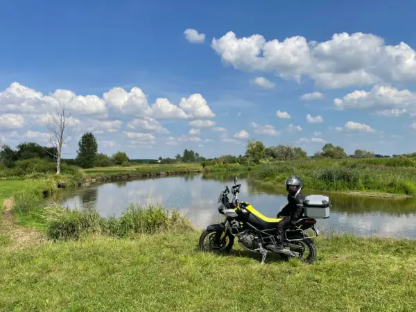 Motocyklista w kasku siedzi na motocyklu obok rzeki, na tle zielonej łąki i błękitnego nieba z chmurami.