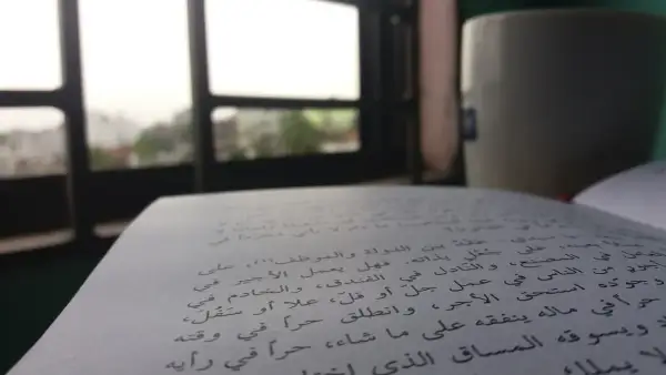 Otwarta księga z tekstem w języku arabskim, obok kubek i widok przez okno.