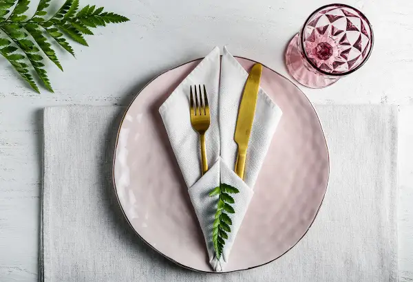 Service de table élégant avec pliage serviette communion, assiette rose, couverts dorés et verre à facettes.