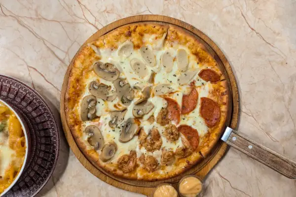 Pizza quattro stagioni avec champignons, saucisses, pepperoni et poulet frit. Une part de pâtes gratinées est visible à gauche.