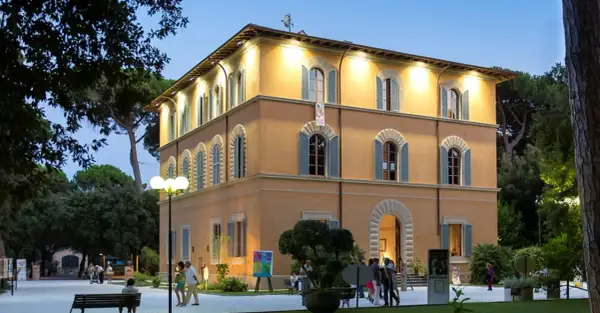 Edificio giallo illuminato nel parco della Versiliana, con persone che passeggiano e un'opera d'arte esposta.