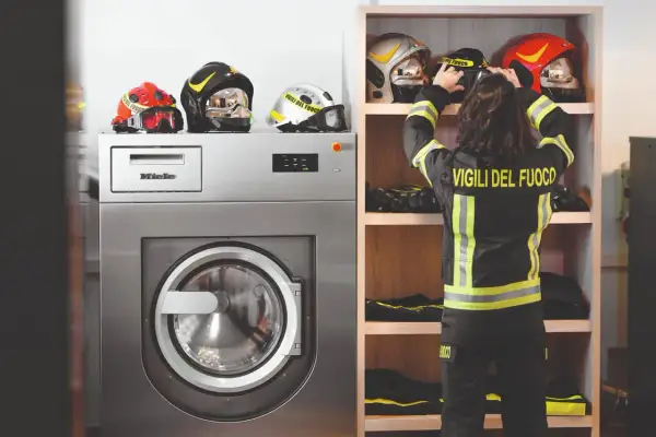 Vigile del Fuoco sistema i caschi accanto a una lavatrice industriale, pronta per il lavaggio abiti da lavoro.
