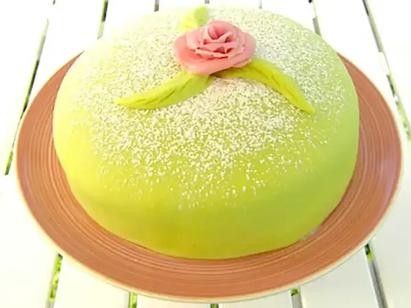 En vacker prinsesstårta med grön marsipan, pudrad med florsocker och dekorerad med en rosa ros. Perfekt för ett prinsesstårta recept!