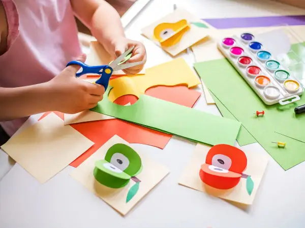 Manos pequeñas cortando papel para manualidades para niños de 3 a 5 años. Pinturas, tijeras y frutas de papel decoran la mesa.