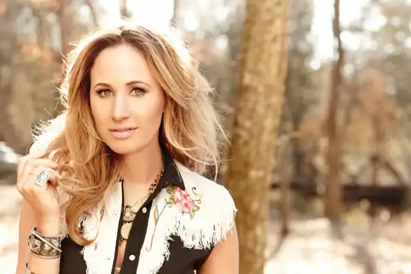 Jessi Alexander, a blonde woman with striking eyes, poses outdoors in a fringed vest adorned with floral embroidery.