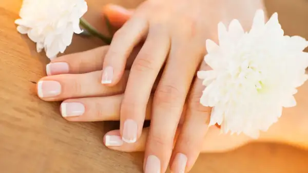 Delikatne dłonie z francuskim manicure, otoczone białymi kwiatami. Paznokcie wyglądają zdrowo, nie są suche.