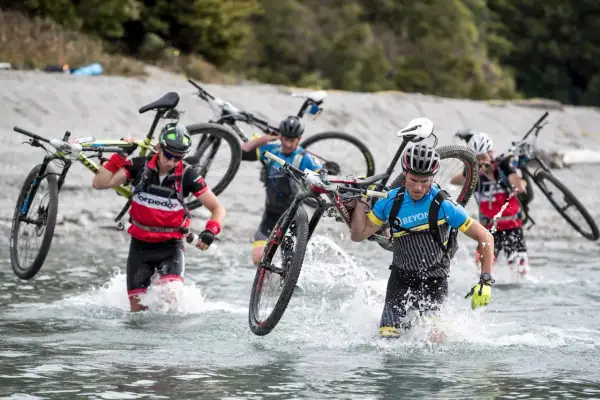 Teilnehmer eines **adventure racing** durchqueren mit Fahrrädern auf den Schultern einen Fluss.