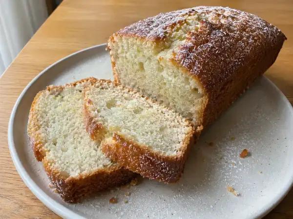 En saftig vegansk sockerkaka, pudrad med florsocker, ligger på en tallrik. Två skivor är upplagda bredvid.