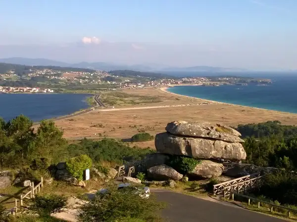 Vista panorámica desde el mirador de Siradella. Rocas gigantescas y un puente de madera en primer plano, con un paisaje de costa, mar azul y un pueblo al fondo.