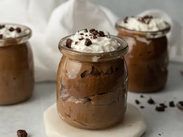 Czekoladowy pudding proteinowy w słoiku, udekorowany bitą śmietaną i kawałkami czekolady.