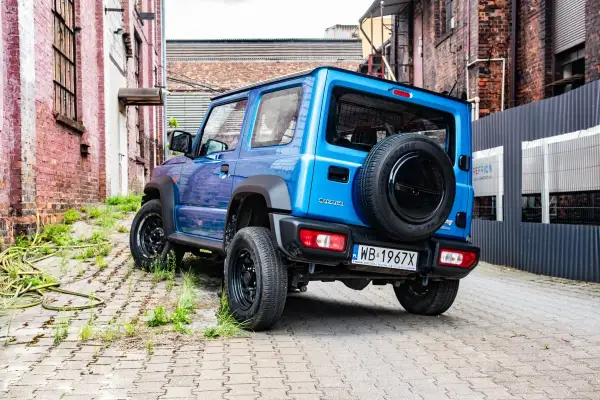 Niebieski Suzuki Jimny z zapasowym kołem na tyle, zaparkowany na brukowanej uliczce między ceglanymi budynkami.
