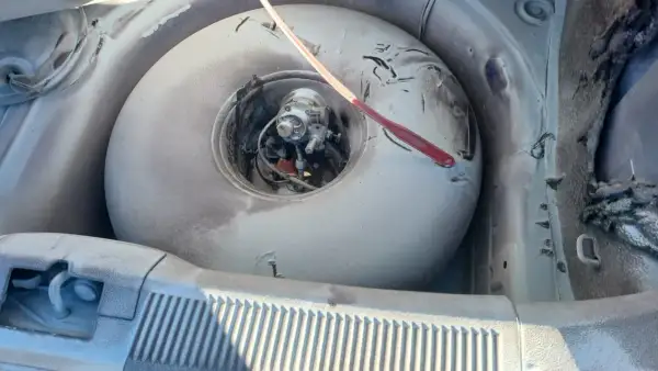 W bagażniku samochodu widać okrągłą butlę LPG, która wygląda na starą i zakurzoną. Czerwony przedmiot leży na jej powierzchni. Czas na wymianę, bo kończy się ważność butli LPG.