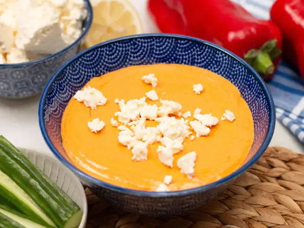 Cremiger Paprika-Feta-Dip in blauer Schüssel, garniert mit Feta-Käse. Dazu Gurkensticks und rote Paprika.