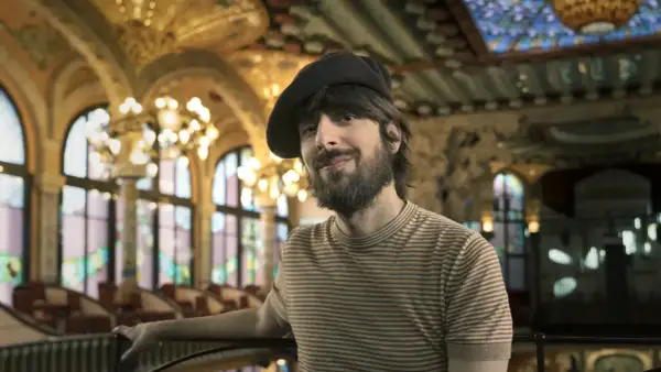 David Ucles sonríe con un gorro y una camiseta a rayas, en un salón con vidrieras y lámparas doradas.