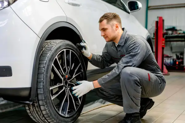 Mécanicien en combinaison grise, avec des gants, examine la roue d'une voiture blanche. Il semble chercher comment dégripper un étrier de frein sans démonter.
