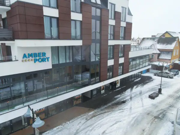 Nowoczesny hotel Amber Port w zimowej scenerii. Na fasadzie widoczny napis "AMBER PORT" i gwiazdki. Ulica pokryta śniegiem.