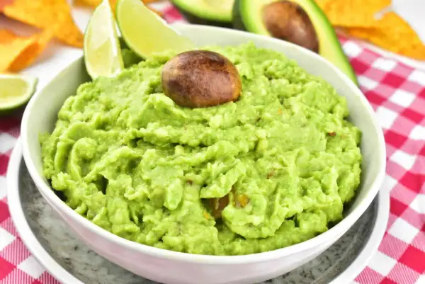 Miseczka guacamole z pestką, limonką i nachos w tle na czerwono-białej kratce.
