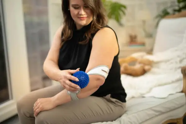Frau massiert ihren Unterarm mit einem blauen Igelball und trägt eine Bandage.