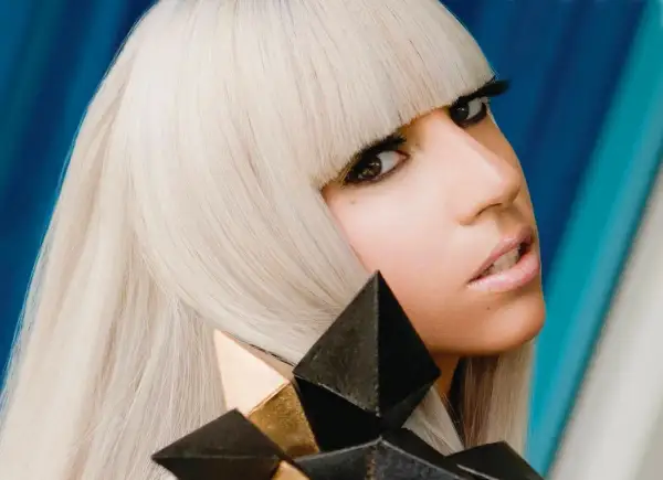 Lady Gaga, con capelli biondo platino e un look audace, cattura l'essenza del suo **inizio carriera** con un'espressione intensa e un accessorio geometrico.