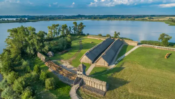Rekonstrukcja osady Biskupin z drewnianymi domami krytymi strzechą, wałami obronnymi i jeziorem w tle.