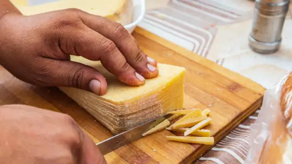 Mano cortando un bloque de queso en finas láminas. La corteza dorada se ve apetitosa, demostrando que se puede comer la corteza del queso.
