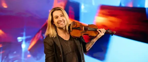 Ein lächelnder Mann mit langen blonden Haaren hält eine Geige. Er könnte der beste Geiger der Welt sein, mit seiner Leidenschaft für Musik.