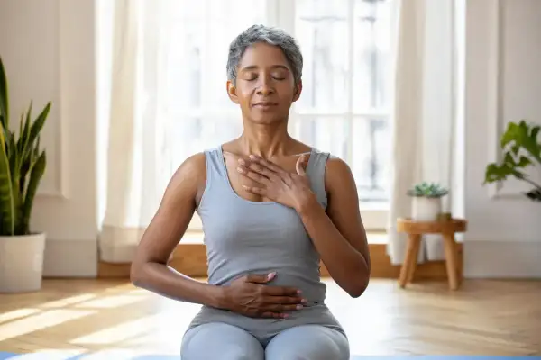 Femme pratiquant une technique de relaxation mentale, mains sur la poitrine et le ventre, yeux fermés, dans une pièce lumineuse.