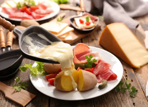 Roztopiony ser raclette spływa na ziemniaki i wędliny. Obok kawałek sera i talerz z dodatkami.