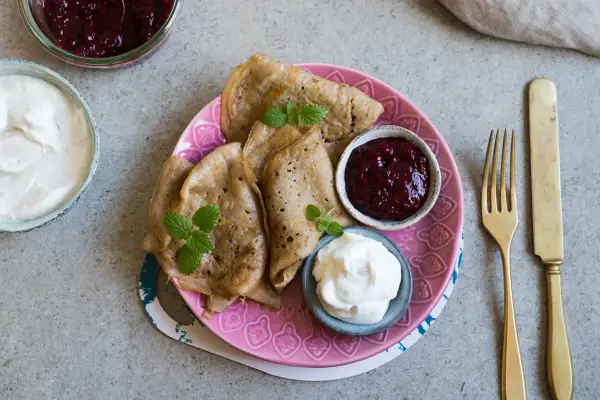 En tallrik med glutenfria pannkakor, serverade med sylt och grädde. En gyllene gaffel och kniv ligger bredvid.