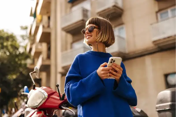 Frau mit blondem Bob, Sonnenbrille und blauem Pullover, die auf ihrem Handy scrollt. Ein rotes Moped steht daneben. Ein toller Look, wie man einen blauen Pullover kombinieren kann, Herren-Style inspiriert.