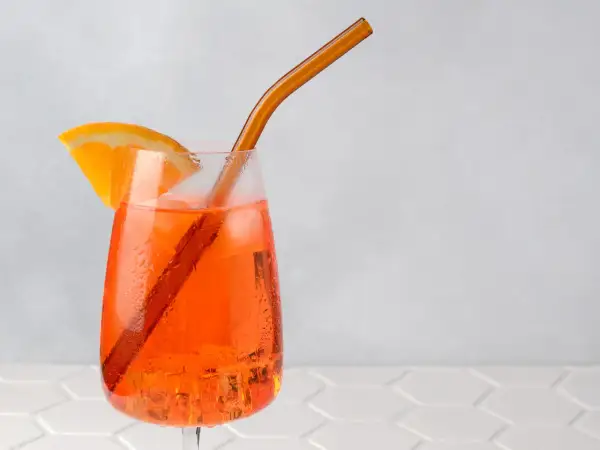 Ein Aperol Spritz mit Orangenscheibe und braunem Strohhalm auf Eis.