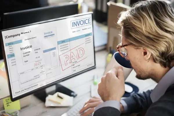 Mężczyzna pije kawę, patrząc na ekran komputera z fakturą z pieczątką "PAID" i informacją o podatku VAT.