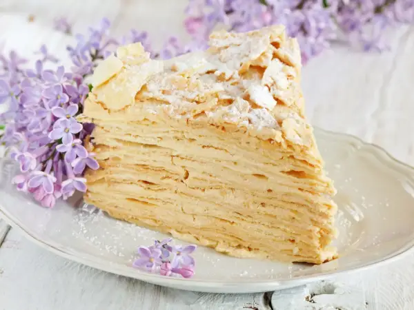 Pyszny tort napoleon z delikatnym kremem i chrupiącymi warstwami ciasta, ozdobiony płatkami migdałów i cukrem pudrem, w otoczeniu kwitnącego bzu.