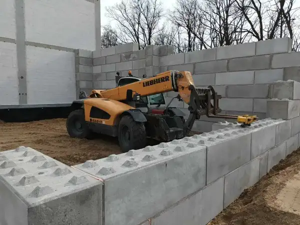 Żółty ładowacz teleskopowy Liebherr układa betonowe bloki, budując mur oporowy.