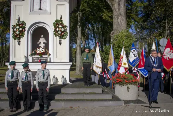 Uroczystość przy kapliczce. Czy imię Bruno jest święte? Dzieci w harcerskich mundurkach i dorosły z łańcuchem na szyi stoją przed kapliczką z figurą i flagami.