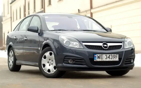 Ciemnoszary Opel Vectra C z polskimi tablicami WE 3439F. Widać jego przednie reflektory, atrapę chłodnicy z logo Opla i zderzak.