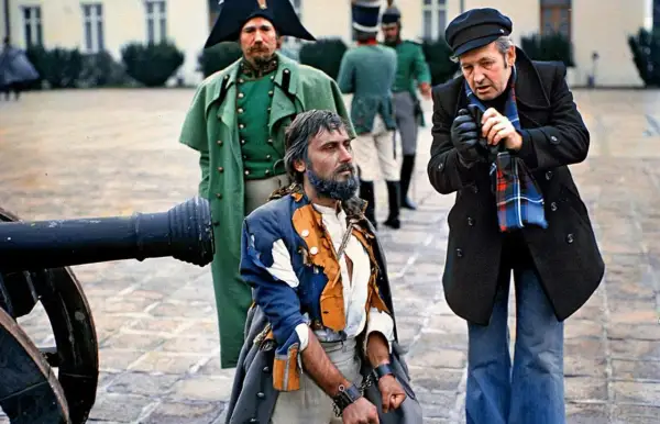 Scena z planu filmowego w polska lat 80. Uwięziony mężczyzna w zniszczonym stroju, obok żołnierze i reżyser.