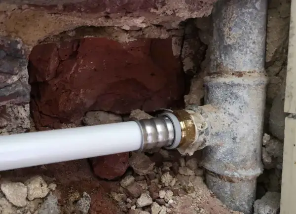 Un nouveau tuyau blanc est raccordé à un ancien tuyau de plomb pour l'eau. La maçonnerie est visible autour des canalisations.