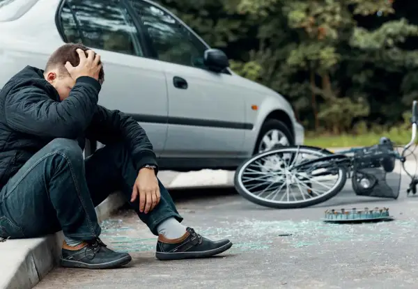 Man zit op stoeprand, hand in haar, na een fietser aangereden te zijn. Fiets ligt omgevallen, met glasscherven op straat.