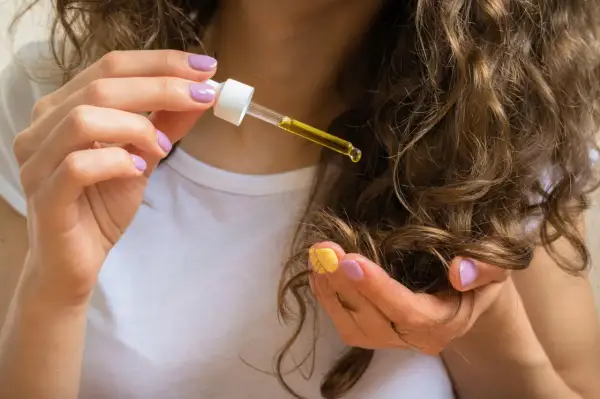 Eine Hand mit lila und gelbem Nagellack hält eine Pipette mit goldenem Ricinus Communis Seed Oil über lockiges Haar.
