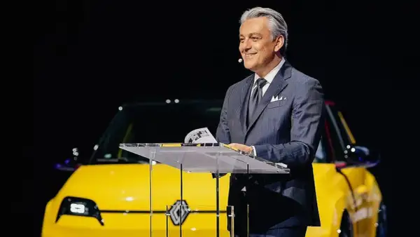 Hombre sonriente en traje, presentando un coche amarillo eléctrico, quizás un nuevo modelo de Volvo Renault.