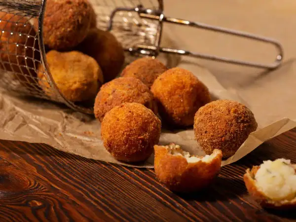Croquetas de patata y pollo doradas y crujientes, algunas en una cesta metálica, otras sobre papel de estraza.