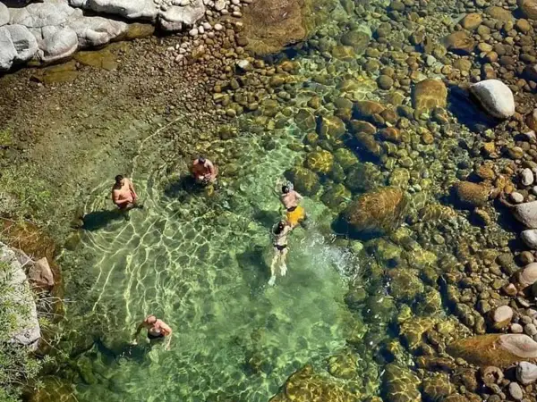 Gente se baña en un río cristalino con fondo de piedras, cerca de un **ponton de la oliva**.