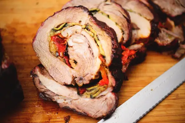 Coppa di maiale allo spiedo, succulenta e ripiena di verdure, pronta per essere affettata su un tagliere di legno.