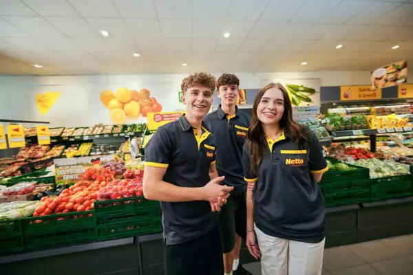 Drei junge Mitarbeiter lächeln in einem Supermarkt. Sie tragen Arbeitskleidung mit dem Netto-Logo. Die Ausbildung zum Elektroniker bietet ein gutes Gehalt netto.