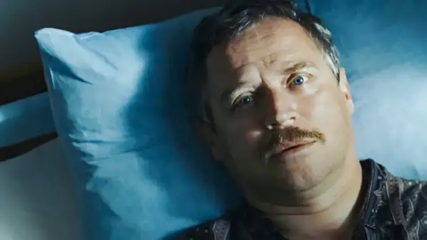 Mężczyzna z wąsami leży na niebieskiej poduszce, jego oczy patrzą w górę. Może to scena z filmu "Dzień świra", gdzie reżyser ukazuje codzienne zmagania bohatera.
