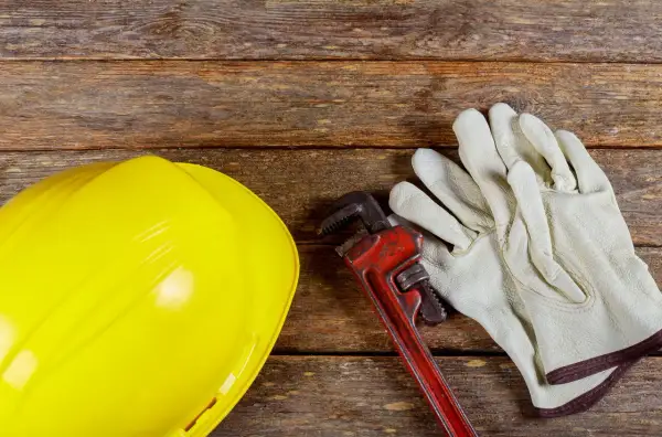 Żółty kask, klucz hydrauliczny i rękawice na drewnianym tle. Zastanawiasz się, dlaczego woda schodzi, a jest bulgotanie w rurach? Czas na fachowca!